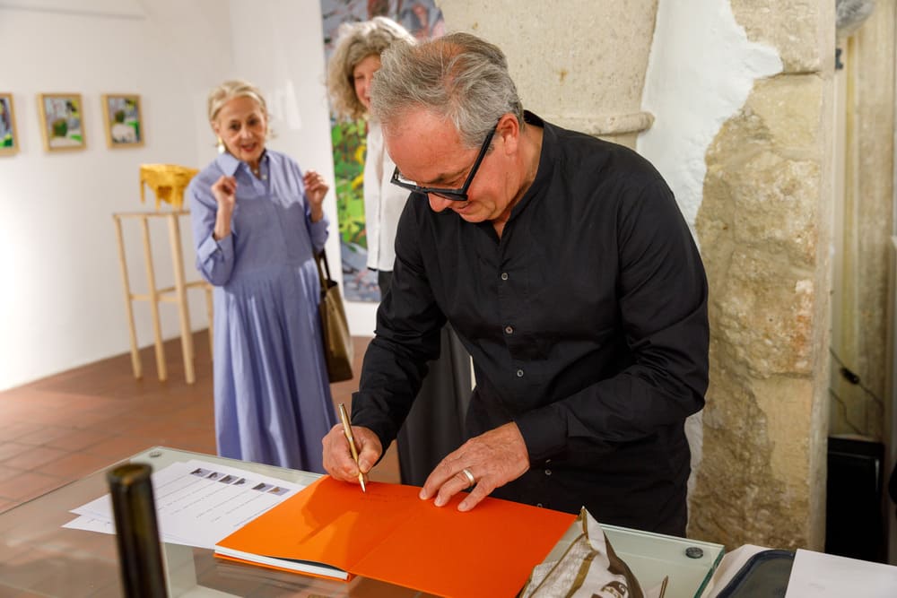 L'artista Giuseppe Donnaloia sorride mentre si appresta a firmare un libro d'arte, circondato dai visitatori all'interno della galleria.