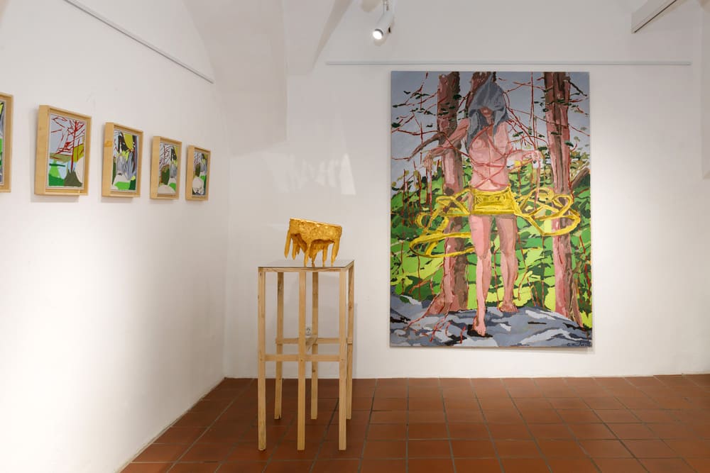 Scorcio espositivo che mostra una scultura in ceramica dorata su un piedistallo e, sulla parete destra, un grande dipinto verticale raffigurante una figura umana immersa in un bosco stilizzato dai colori vivaci.