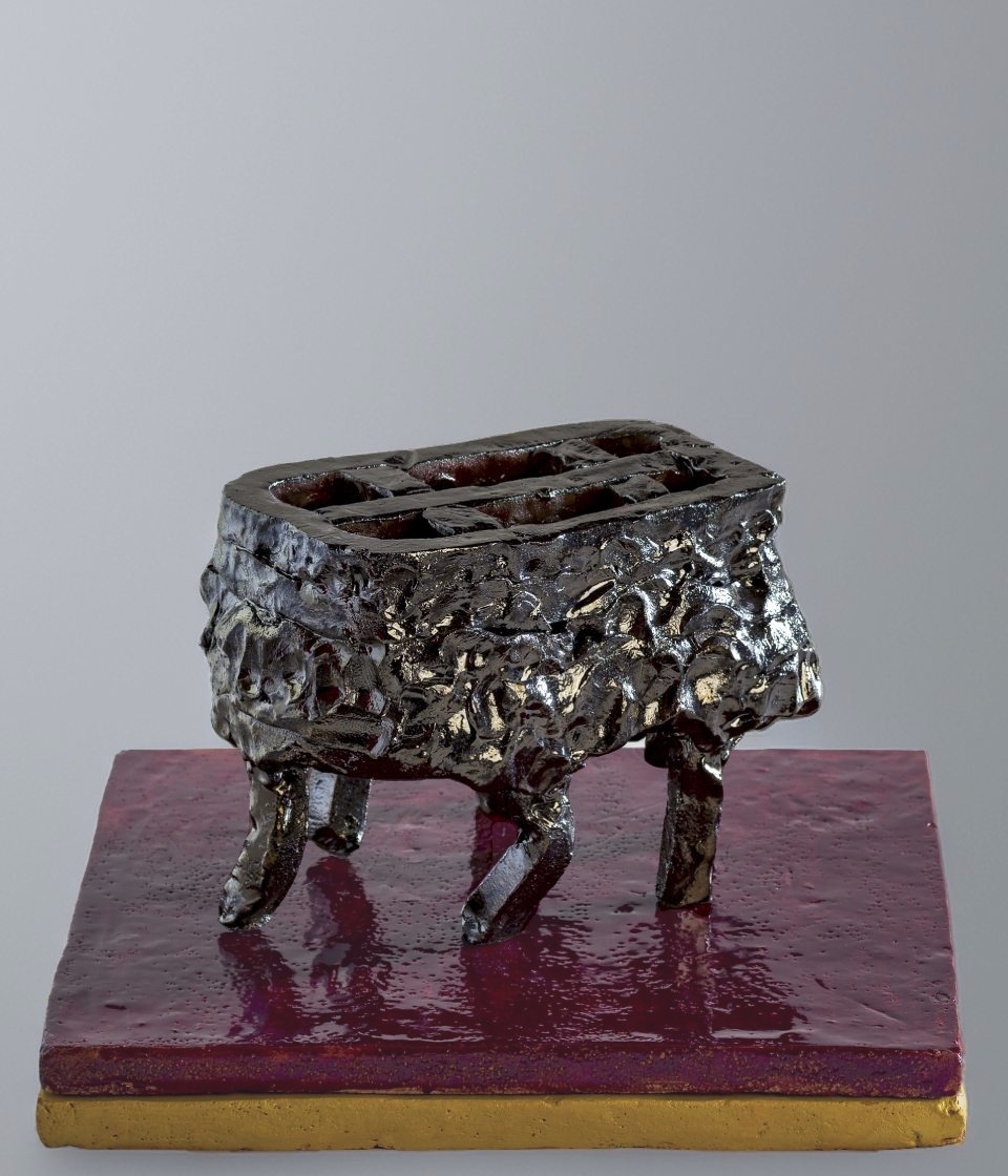 Scultura in ceramica nera con riflessi argentei e metallici, dalla forma di un contenitore rettangolare con pareti spesse e irregolari. L'opera sembra fluttuare sopra una base bordeaux grazie a gambe asimmetriche che suggeriscono un equilibrio precario.