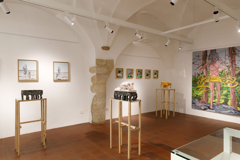 Una veduta panoramica dell'allestimento nella Galerie Panta Rhei; le sculture in ceramica della serie "In bilico" sono esposte su alti piedistalli in legno chiaro, circondate da tele di varie dimensioni sotto le volte bianche della galleria.