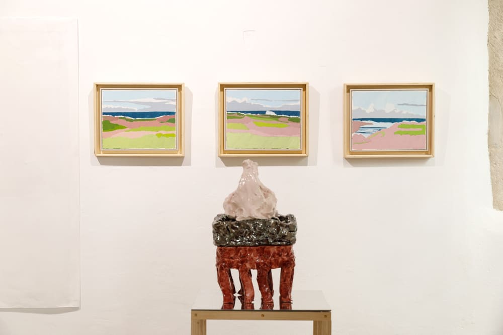 Primo piano frontale di una scultura in ceramica bordeaux e rosa poggiata su una superficie specchiante; sullo sfondo, tre dipinti incorniciati che raffigurano paesaggi costieri stilizzati con tonalità rosa, verdi e blu.