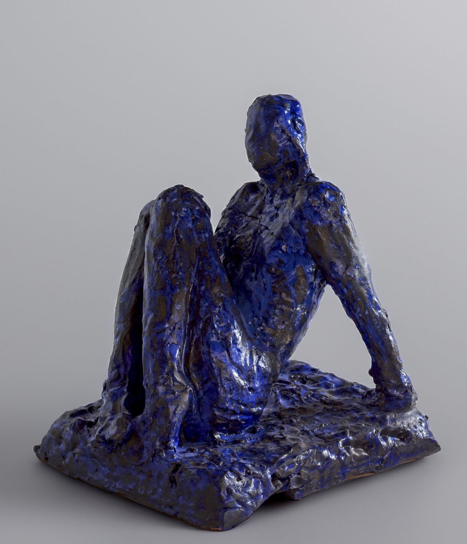 Scultura figurativa in ceramica che rappresenta una figura umana seduta in una posa raccolta, con le ginocchia sollevate e le braccia che poggiano a terra dietro la schiena. L'opera è interamente rivestita da uno smalto blu cobalto vibrante e lucido, steso su una superficie fortemente strutturata e non levigata che lascia intravedere il corpo sottostante.